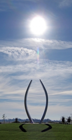 The sun in a cloudy blue sky appears directly above Beginnings sculpture on grassy green landscape.