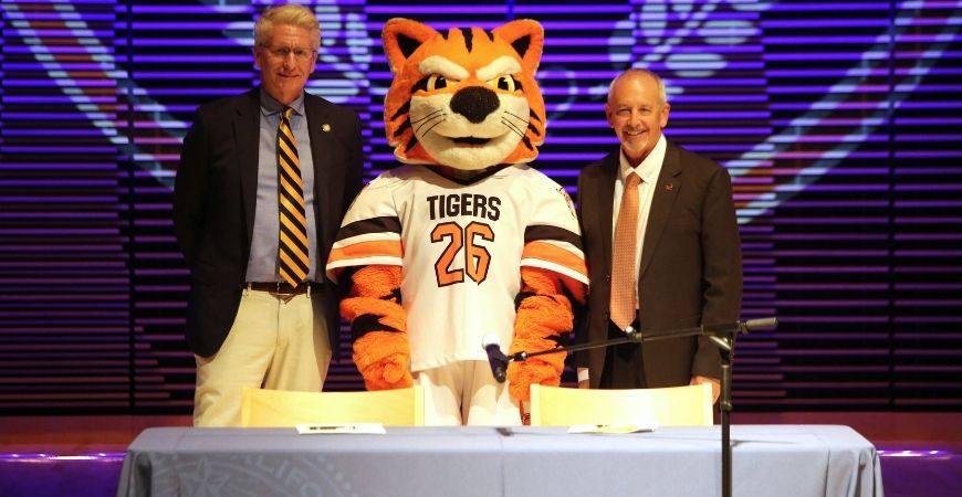 Two men in suits stand beside a tiger mascot wearing a white “TIGERS 26” jersey behind a table on a stage with purple lighting.