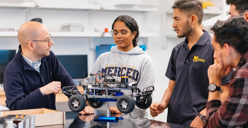 Image depicts UC Merced mechanical engineering Professor Ricardo Pinto de Castro talking to students near a small robot.