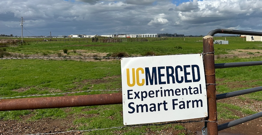 Depicted is a sign reading "UC Merced Experimental Smart Farm" with the university campus in the background.