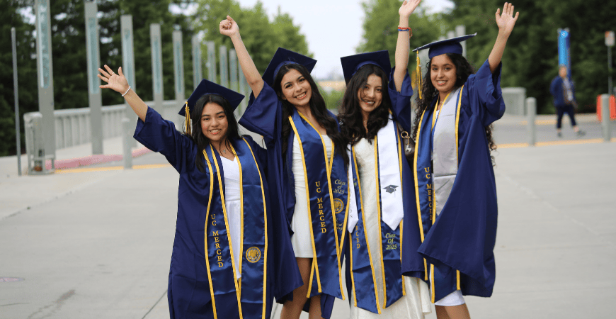 Four female UC Merced graduates in cap and gown pose in a celebratory fashion