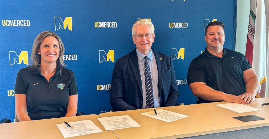 Three people sit behind a table in front of a blue UC Merced backdrop, smiling for a photo after signing documents.