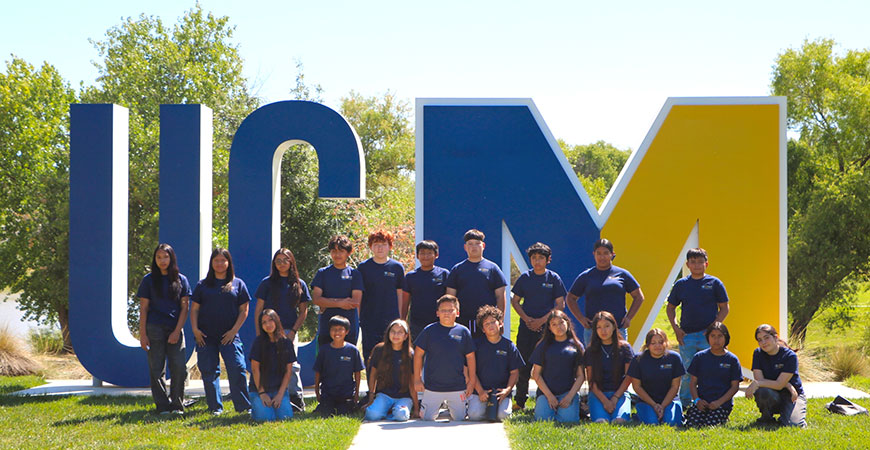 Depicted are two two lines of middle-school students standing in front of the blue and gold UCM on the UC Merced campus.
