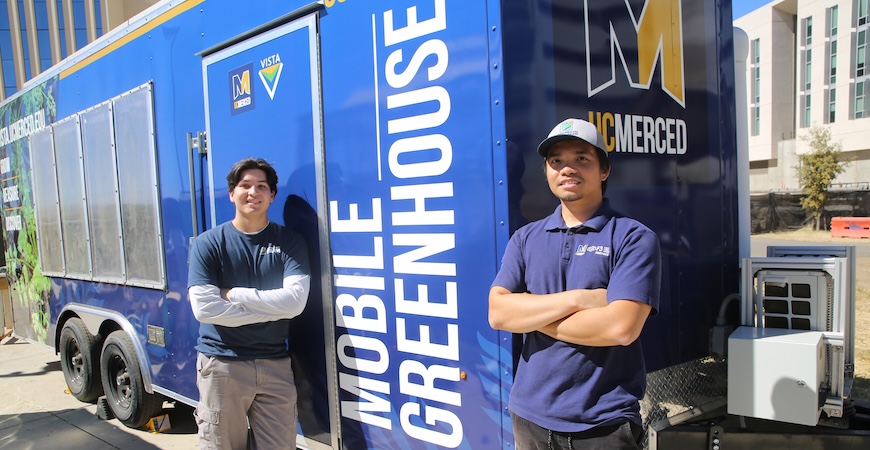 Specialists Isaac Leyva and Ethan Vu are pictured standing outside the blue trailer with white lettering reading "Mobile Greenhouse."