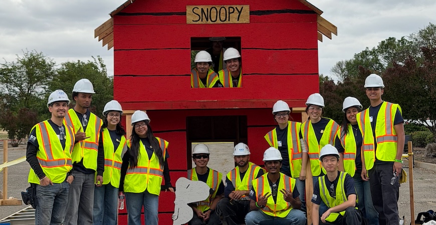 Depicted are students in yellow vests and hard hats standing around and in a two-story Snoopy doghouse.