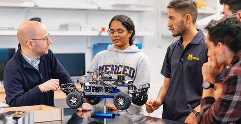 Image depicts UC Merced mechanical engineering Professor Ricardo Pinto de Castro talking to students near a small robot.