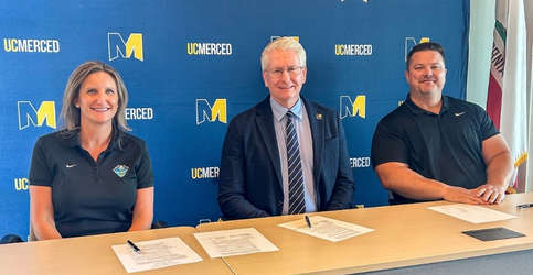 Three people sit behind a table in front of a blue UC Merced backdrop, smiling for a photo after signing documents.