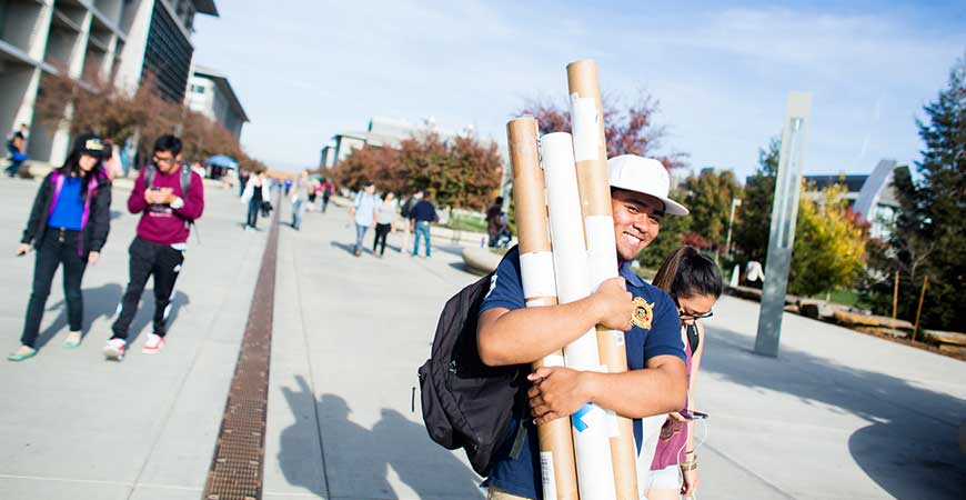 A smiling male student walks along Scholars Lane while carrying an arm full of papers.