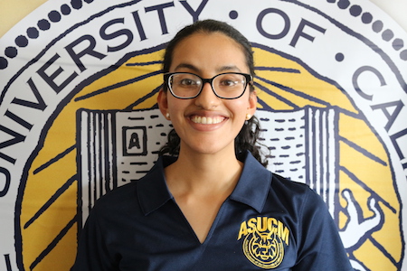 Akhila Yechuri, wearing black-rimmed eyeglasses and a navy blue Associated Students of UC Merced polo shirt, stands in front a University of California seal.