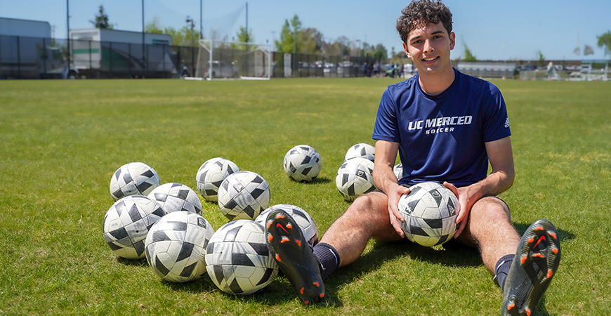 A student poses with several soccer balls on an athletic field.