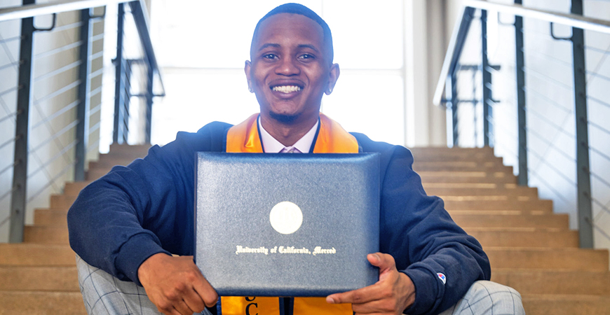 A student sits in a stairwell and smiles while holding a diploma cover.