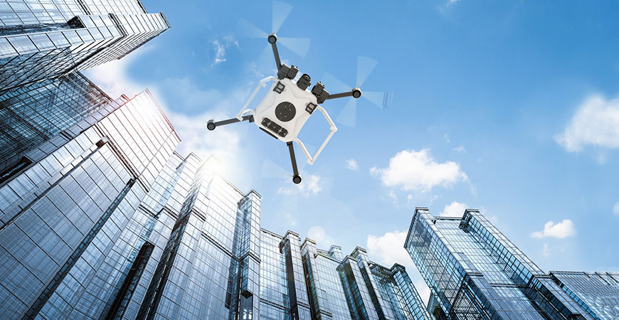 A drone, viewed from below, flies between tall buildings on a partly cloudy day.