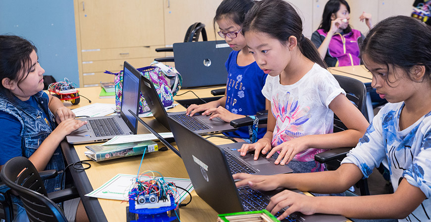 Four female middle school students in summer robotics academy are gathered around a desk. All four students are sitting in front of laptops and writing code for their robots.