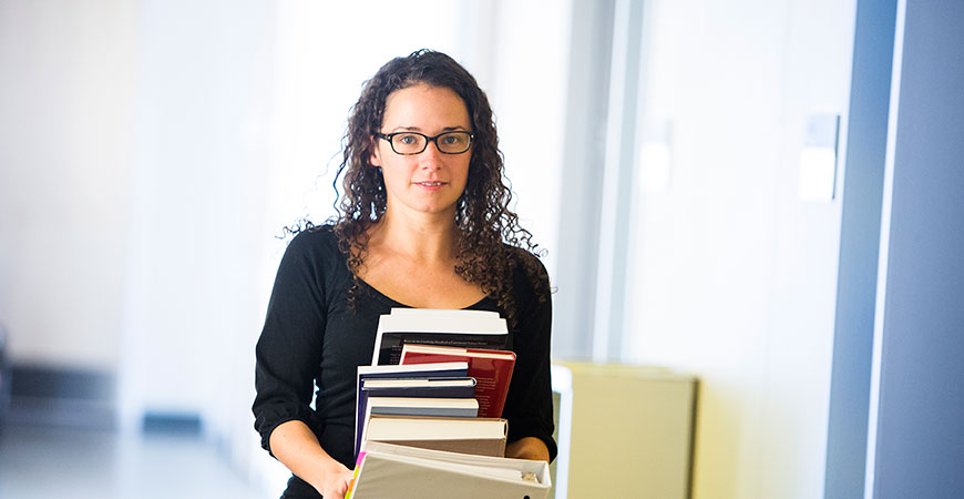 Woman with dark curly hair and glasses carrying a stack of books