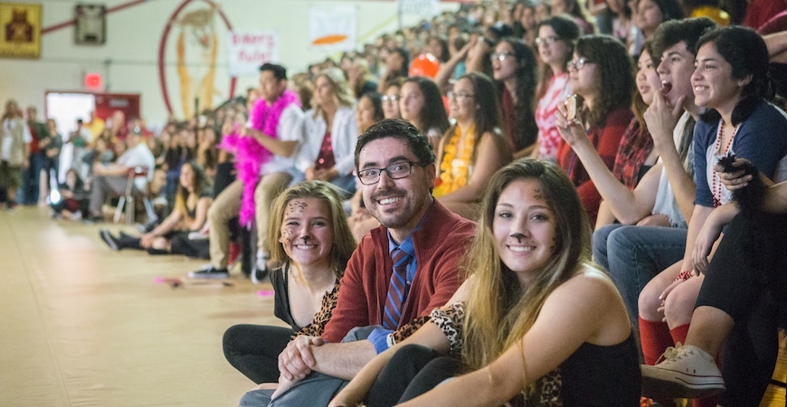 Students and teachers are seated on the floor and bleachers in a high school gymnasium while attending a pep rally in Merced.