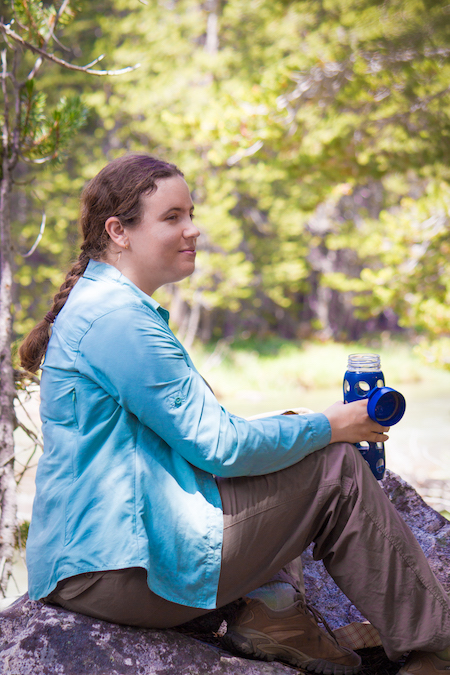 Kaitlin Lubetkin, wearing a turquoise jacket, brown pants, and brown hiking boots, poses atop a rock with trees in the distance behind her.