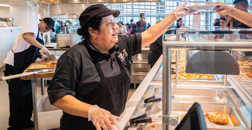 A female food services worker serves a customer while another employee slices pizza in the background.