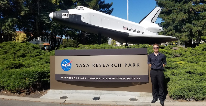 A student stands in front of a sign that reads "NASA Research Park." Above the sign is a model of a NASA space shuttle.