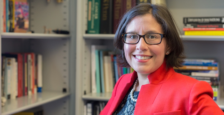Professor Susana Ramirez in a red jacket and black-rimmed glasses stands in front of bookshelves filled with books.