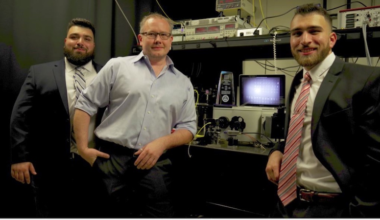 Three men pose in front of scientific equipment.