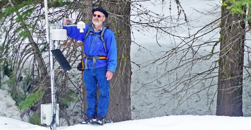 A man in a blue jacket, blue jeans and sunglasses stands in a snowy forest repairing an electronic sensor device.