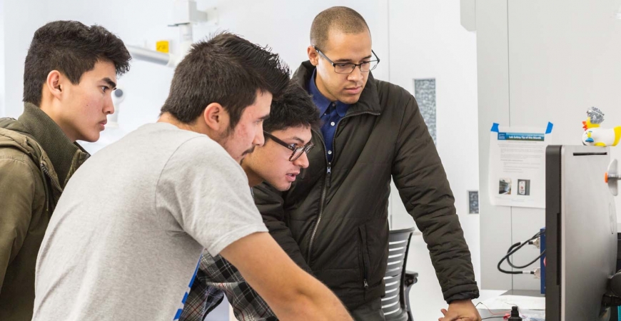 Four male students huddle around a computer as they work.