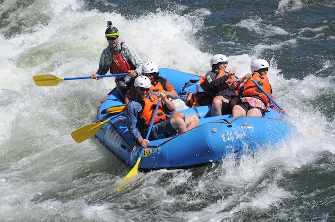 Five people holding oars and wearing orange life vests rafting down a river in a blue raft.