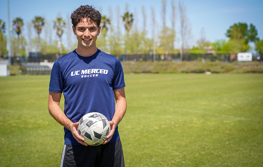 UC Merced student Edwin Casillas poses with a soccer ball on a field.
