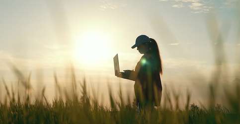 A female farmer is depicted holding a laptop computer in a field with the sun behind her.